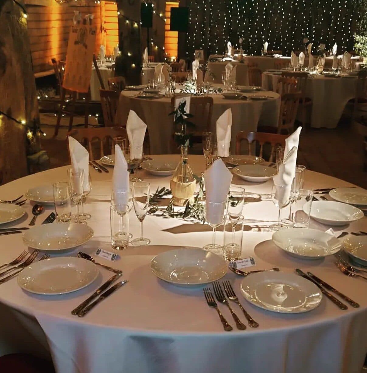 Round table laid with white plates, folded napkins and silver cutlery under fairy lights