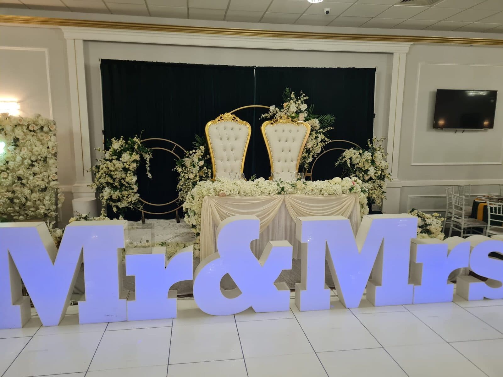 Light-up Mr and Mrs letters in front of gold throne chairs, white flowers and a black backdrop