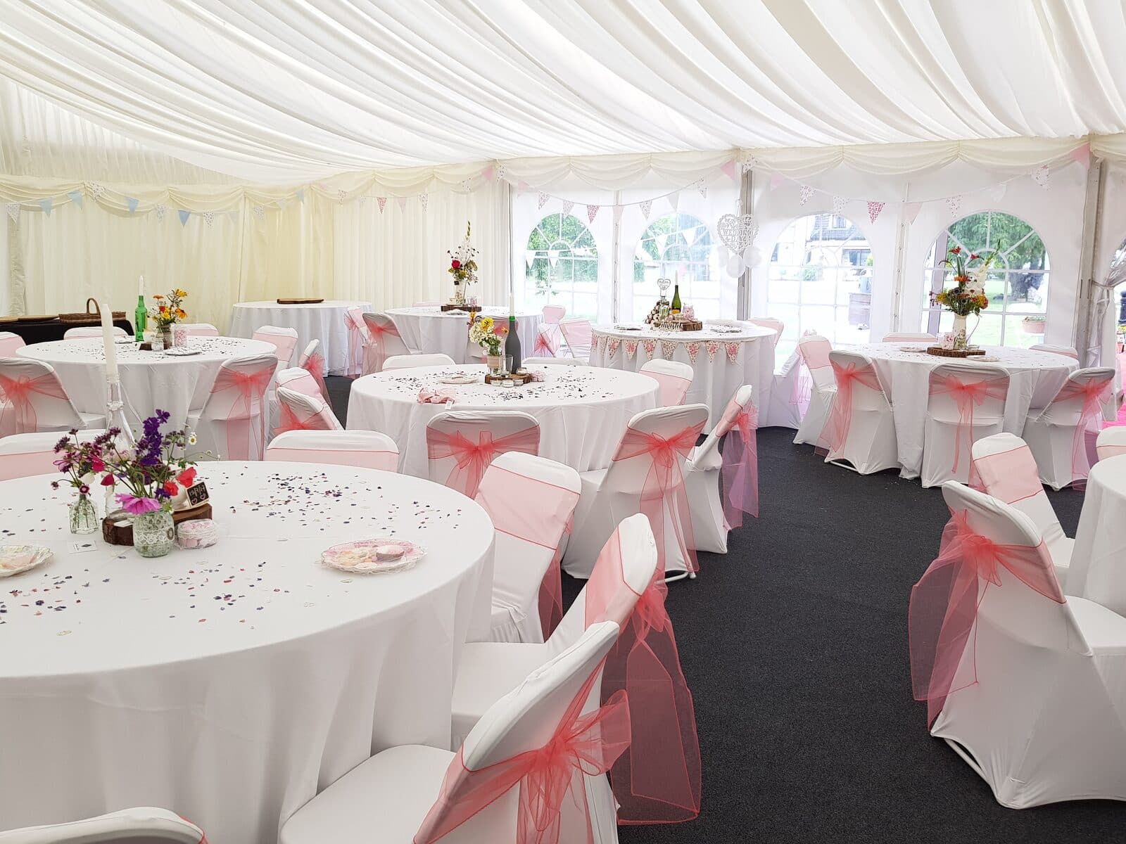 Marquee with white chair covers, red and pink organza sashes and small flower jars on round tables