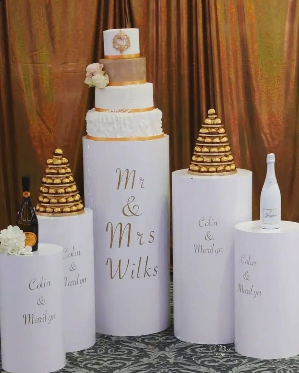 Tiered wedding cake on white cylinder plinths with gold lettering, beside profiterole towers and champagne bottles, in front of a copper curtain