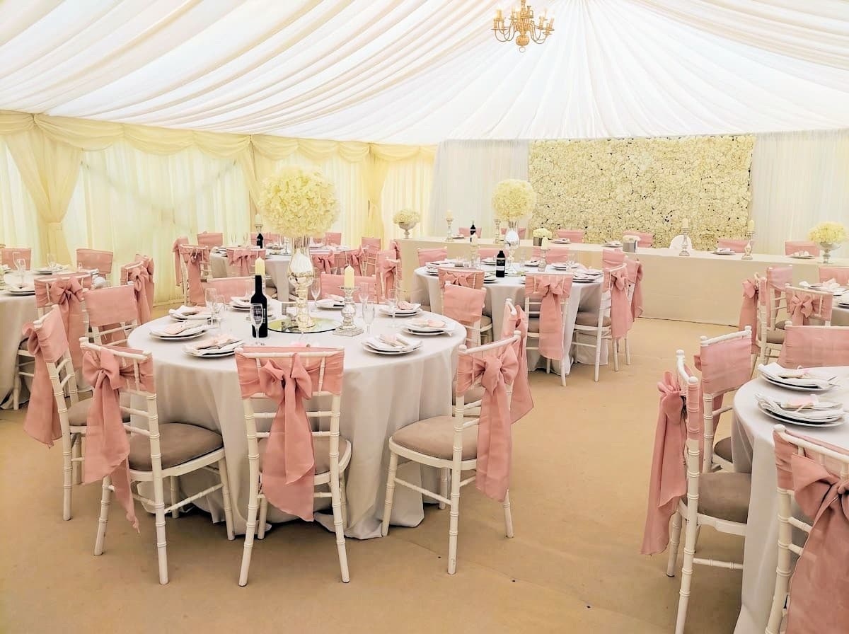 Marquee laid for a wedding: round tables with white chairs and blush pink sashes, white flower centrepieces and a flower wall behind the top table