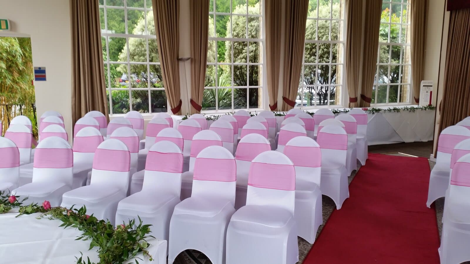Wedding ceremony room set with white chair covers and pink organza sashes, either side of a red aisle carpet